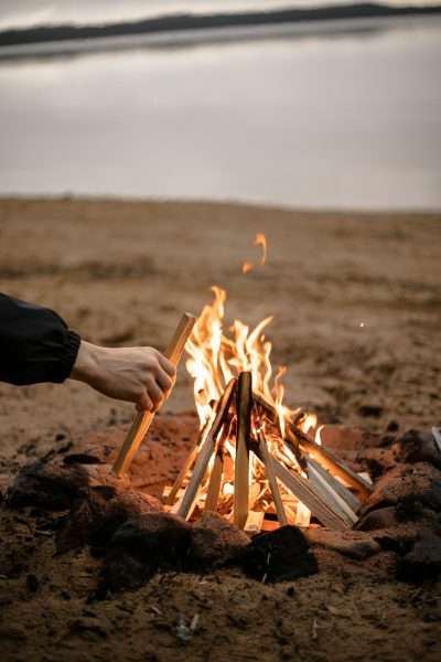 A close-up of a bonfire being tended to on a sandy beach at twilight.